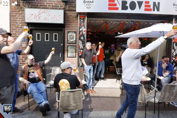 Groep mensen buiten een café, proostend met glazen in de lucht, vrolijke sfeer.
